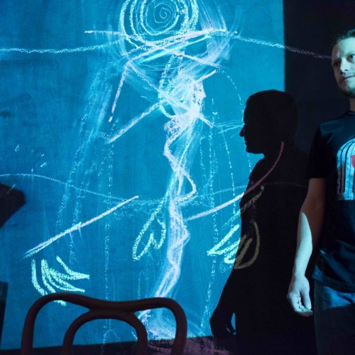 This Stays in the Room: Man standing in front of neon blue projection of hand-drawn squiggles