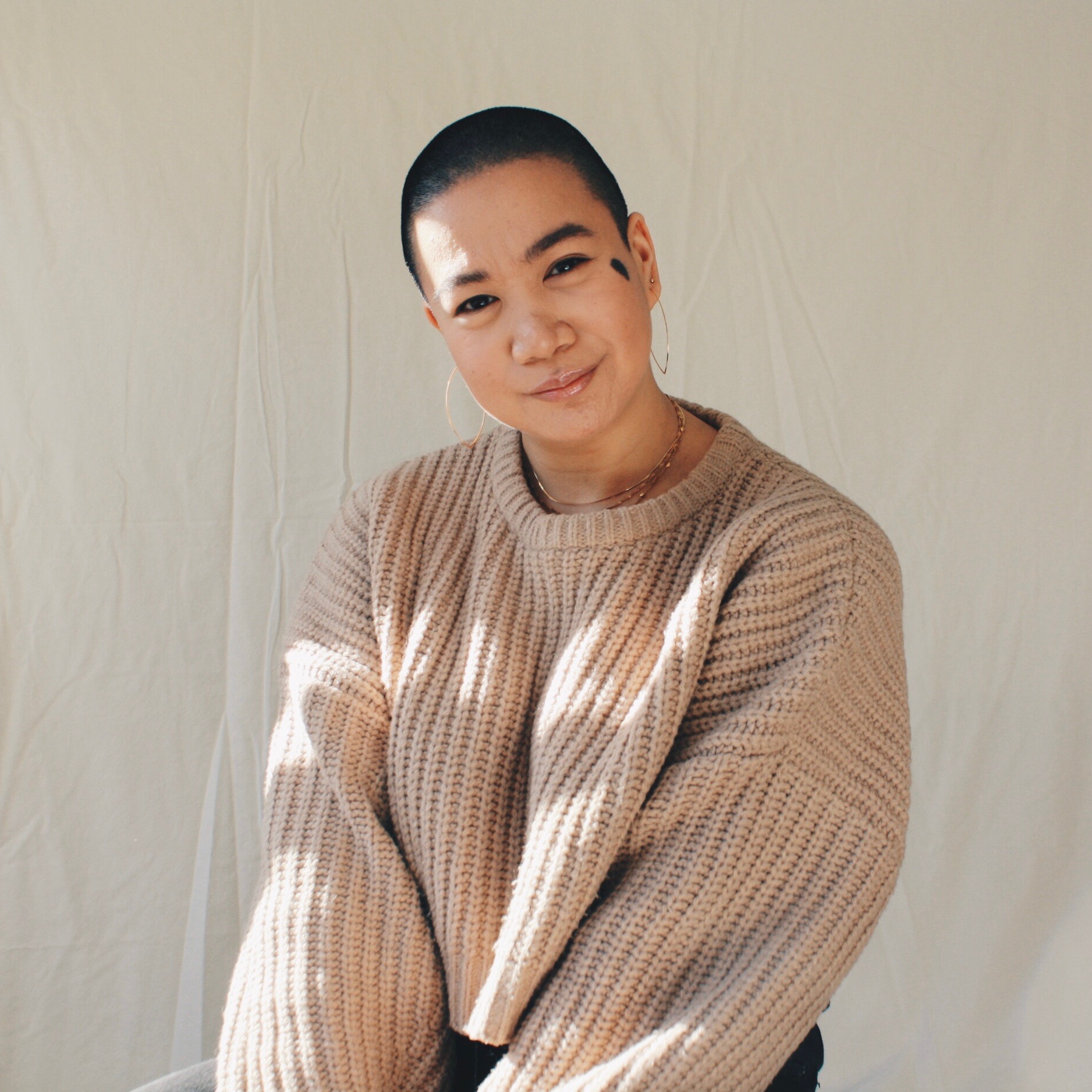 Kamila, a Filipinx woman with a shaved head and an oval birthmark on her left cheek, smiles in front of a white background. Sunshine glints across her face and gold jewellery, creating a mosaic of shadow and light across her light brown sweater.