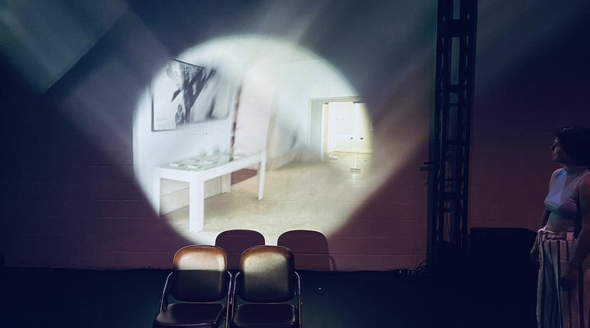 A dark image of a woman looking at the back wall where there is a different room being projected.